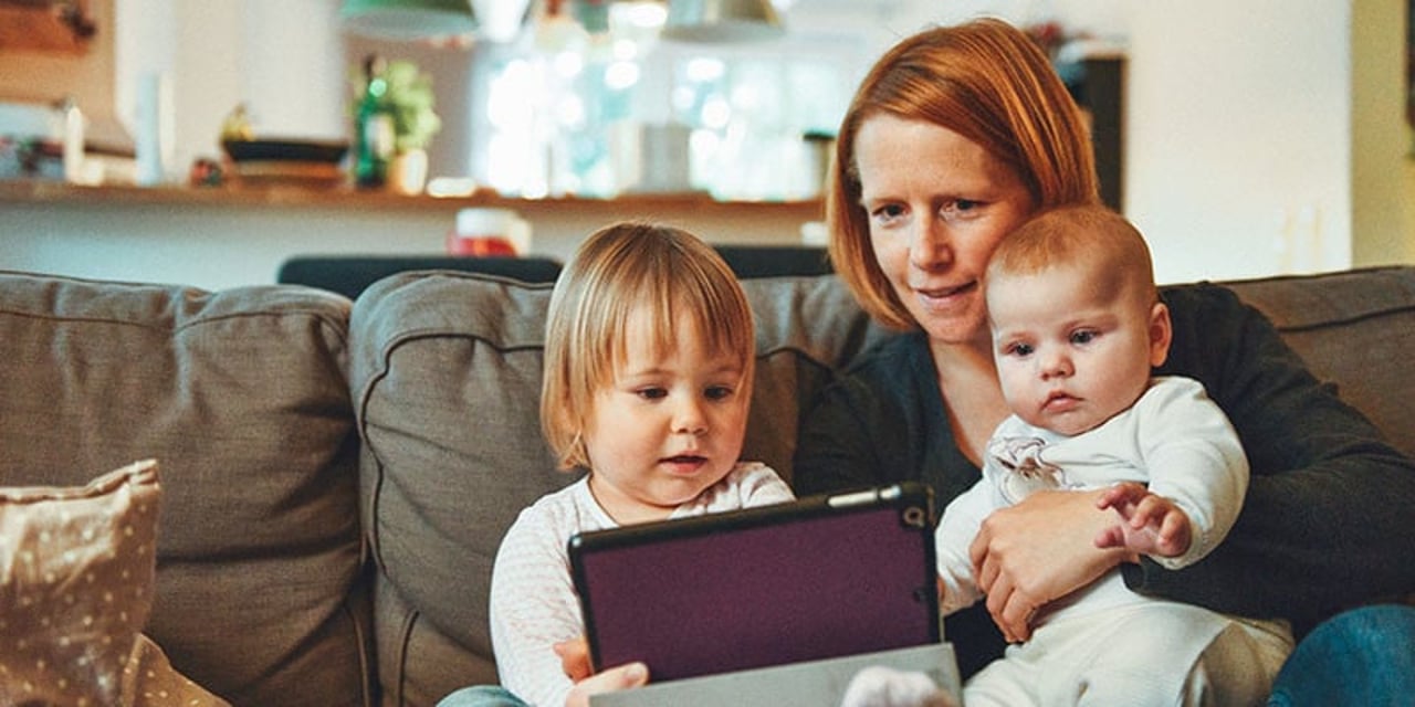 A woman sits on a grey sofa with two children on her lap. They are all looing at a purple tablet.