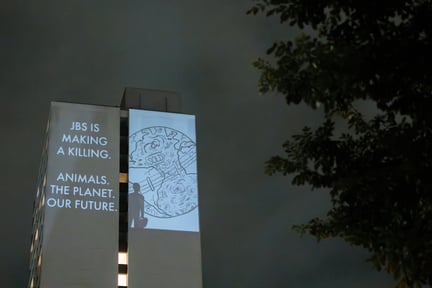 Projection on the side of a tower block saying 'JBS is making a killing. Animals. The Planet. Our future.