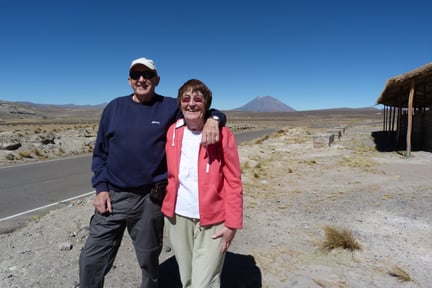 Elderly couple smiling, standing beside a road in a desert landscape with a distant mountain. The sky is clear and blue.