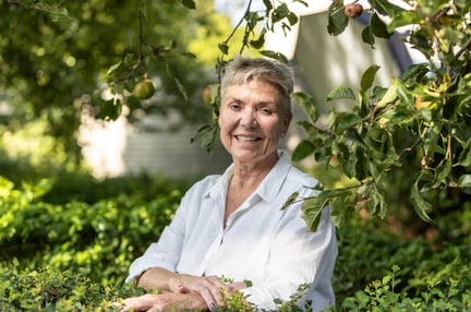 A senior woman with a white blouse and short hair smiling at the camera surrounded by greenery in what seems like a garden