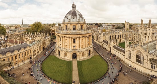 Aerial view of Oxford University with the Radcliffe Camera at centre, surrounded by historic buildings and people running on pathways.