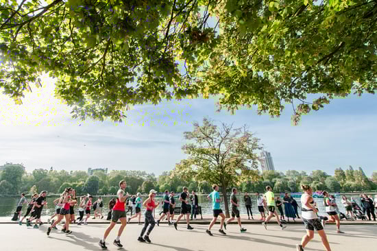 A group of runners jog along a lakeside path under green leafy trees, with a clear sky and a city skyline in the background.