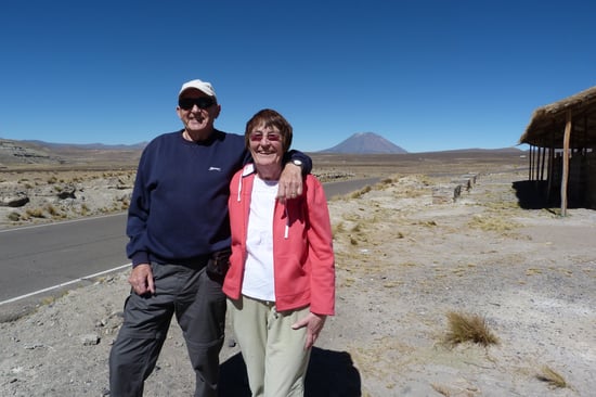 Elderly couple smiling, standing beside a road in a desert landscape with a distant mountain. The sky is clear and blue.