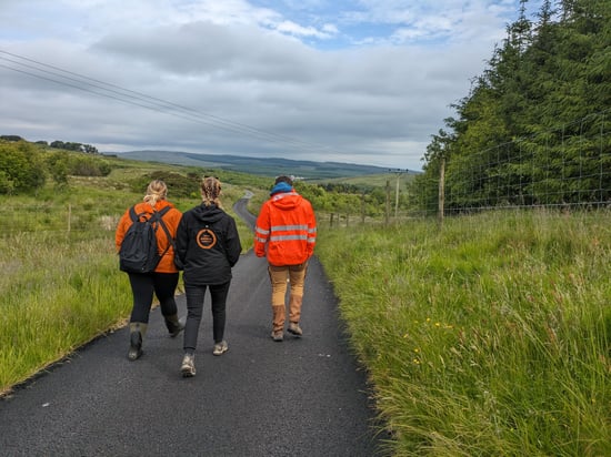 Three people wearing outdoor clothing walk down a rural paved road surrounded by green fields and trees under a cloudy sky.