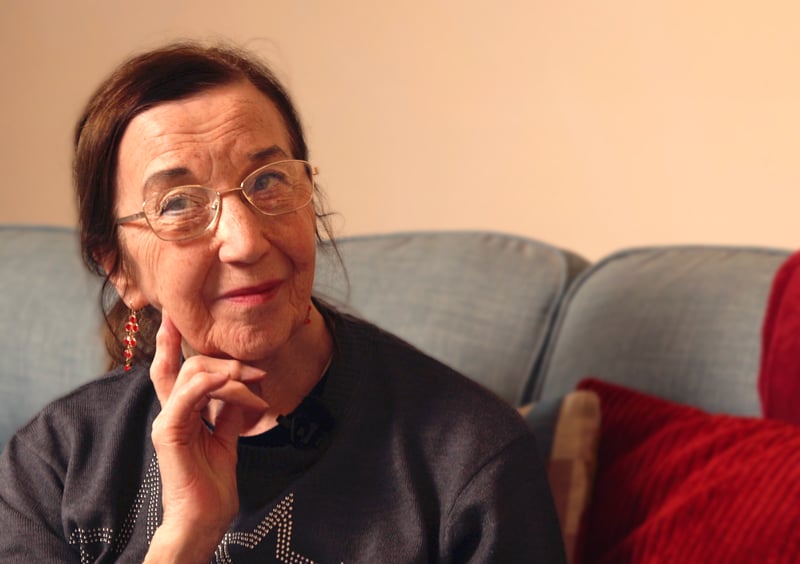 Julie is sitting on her soft posing for the camera. She is wearing glasses and red earrings.